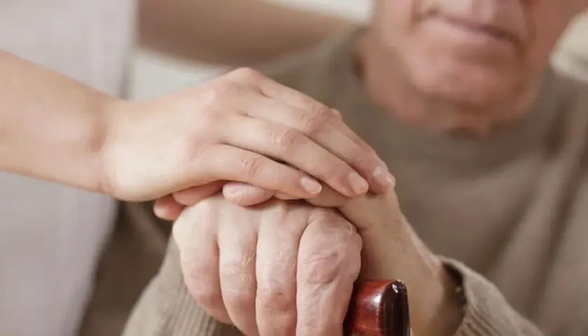 Dłoń opiekunki na dłoni starszego mężczyzny, który trzyma laskę. Wsparcie i troska, jak przy leczeniu np. chorobą Parkinsona, gdzie **Madopar** może pomóc.
