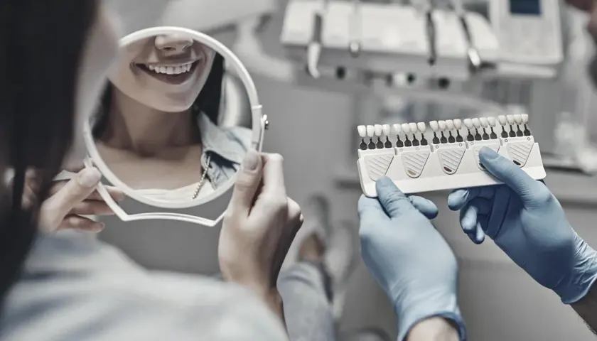 Dentysta w rękawiczkach pokazuje pacjentce wzornik kolorów zębów, pacjentka ogląda swój uśmiech w lustrze.