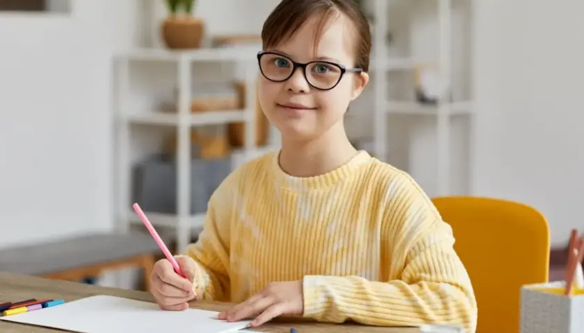 Dziewczynka z trisomią 21, w okularach, z uśmiechem rysuje kredką.