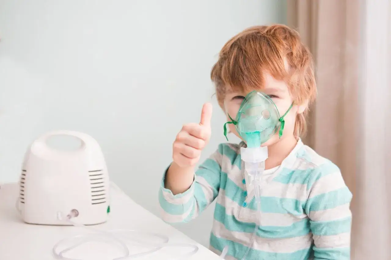Chłopiec z maską inhalacyjną budixon neb daje kciuk w górę, obok stoi biały inhalator.