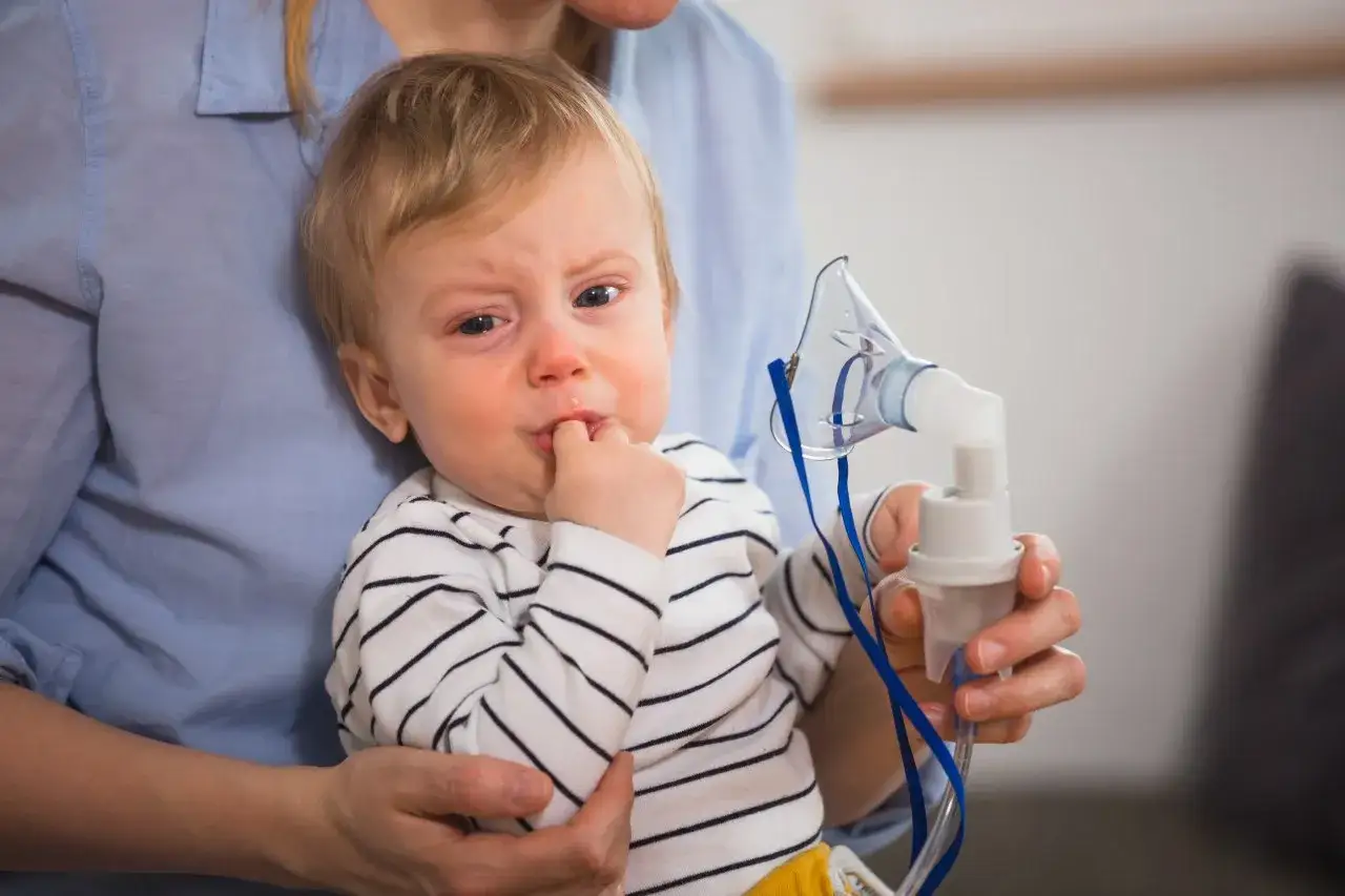 Płaczące dziecko trzymane przez mamę, obok nebulizator z lekiem budixon neb.